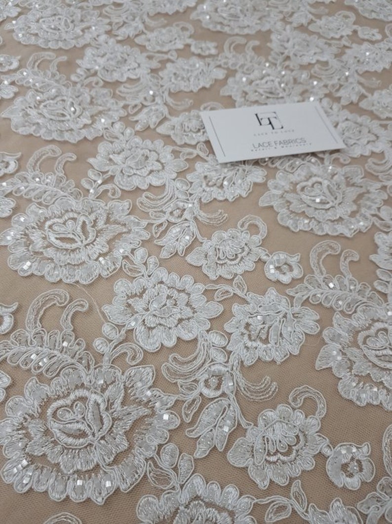 May include: Detailed shot of white floral lace fabric with delicate embroidered patterns. The lace showcases a repeating design of flowers and leaves, adorned with tiny beads. A white card with the text "LACE FABRICS" is on the fabric.