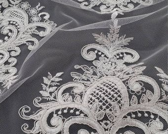 May include: Close-up of white lace fabric with intricate floral and scrollwork embroidery. The delicate design is set against a gray background. A card with the text "LACE TO LOVE" is visible. This fabric is suitable for bridal wear and other special occasion garments.