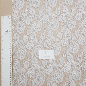 May include: White floral lace fabric with a detailed pattern of flowers and leaves. A ruler with centimetre and inch measurements is visible along the left edge. The fabric is displayed on a neutral background. The text "LACE FABRICS" is visible.