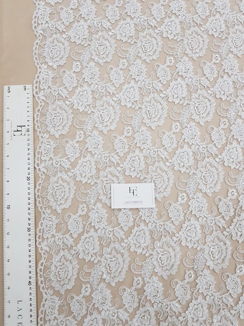 May include: White floral lace fabric with a detailed pattern of flowers and leaves. A ruler with centimetre and inch measurements is visible along the left edge. The fabric is displayed on a neutral background. The text "LACE FABRICS" is visible.