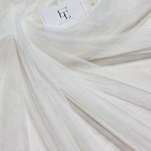 Peut inclure: Tissu de tulle blanc avec une texture délicate et transparente. Le tissu est drapé et plié, créant des ombres et des reflets doux. Une petite carte avec le texte "LACE TO LOVE" est visible.
