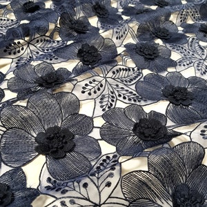 May include: Close-up of navy blue floral lace fabric. The fabric features embroidered flowers with raised, three-dimensional centers. The design includes detailed leaves and stems, creating a textured and elegant appearance. The lace is set against a light background.