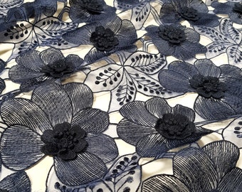 May include: Close-up of navy blue floral lace fabric. The fabric features embroidered flowers with raised, three-dimensional centres. The design includes detailed leaves and stems, creating a textured and elegant appearance. The lace is set against a light background.