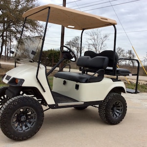 May include: A white golf cart with a tan canopy, black seats, and black wheels with a gray rim. The cart has a windshield and a rearview mirror.