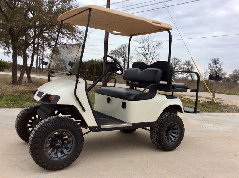 May include: A white golf cart with a tan canopy, black seats, and black wheels with a gray rim. The cart has a windshield and a rearview mirror.
