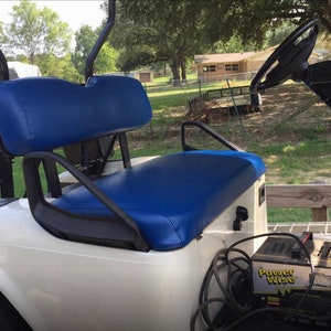 May include: White golf cart with a blue seat and a black steering wheel. The cart has a black battery charger labeled "Power Wise" attached to the back.