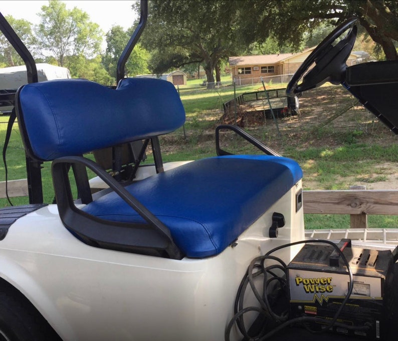 May include: White golf cart with a blue seat and a black steering wheel. The cart has a black battery charger labeled "Power Wise" attached to the back.