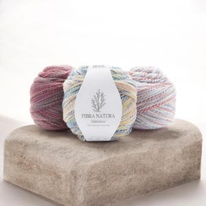 May include: Three round balls of Fibra Natura Cobblestone yarn. The left ball is a deep mauve, the center ball is a mix of light blue, yellow, and white, and the right ball features a blend of light blue, pink, and white variegated colors.
