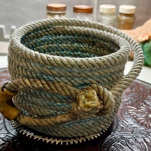 May include: A handcrafted rope basket with a handle, featuring a mix of beige, blue, and brown tones. The basket has a unique knot detail and a leather accent. The design is rustic and textured, suggesting a Western or nautical theme.
