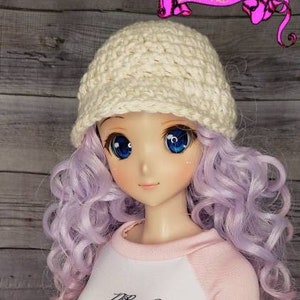 May include: A doll wearing a white crocheted hat, a white and pink long-sleeved shirt with the text "Dream Couture" on the front, and black pants.
