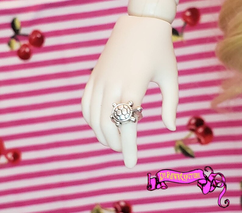 May include: A silver turtle ring with a detailed shell design. The ring is worn on a hand with a pink and white striped background.