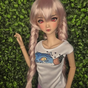 May include: A doll with pink hair styled in braids, red eyes, and a white t-shirt featuring a blue cat graphic. The doll is wearing purple pants and has a piercing. The background is a wall of green foliage.