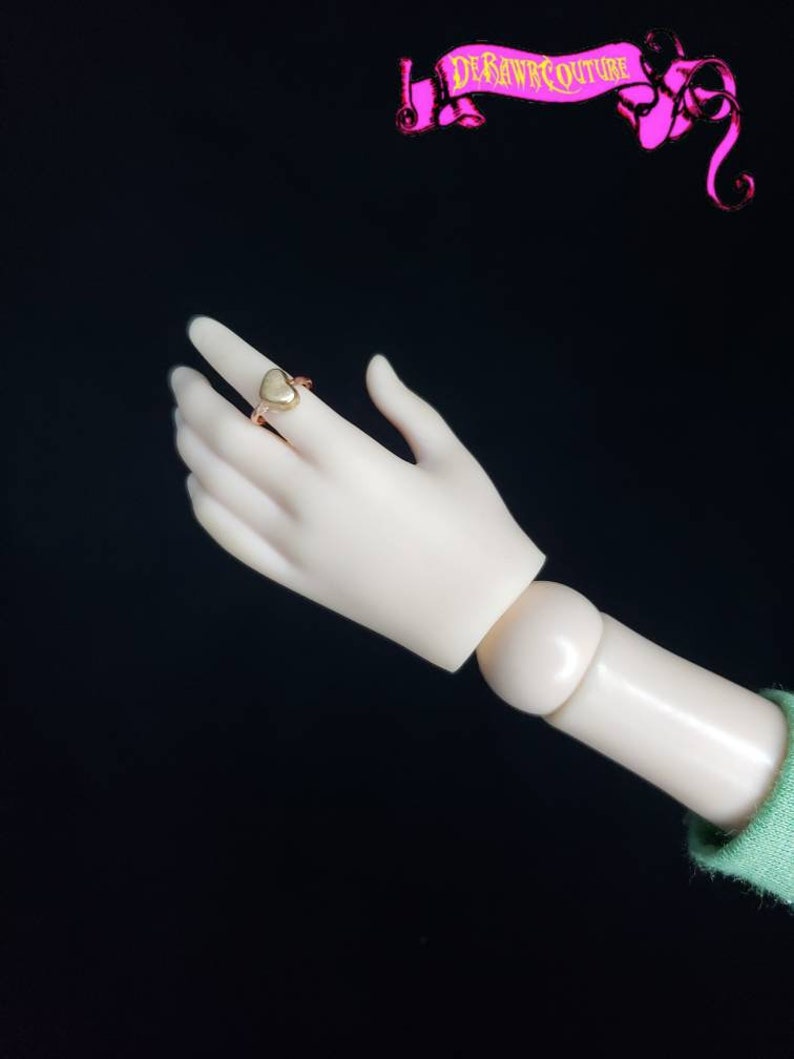 May include: A gold heart-shaped ring with a smooth, polished finish. The ring is worn on the index finger of a hand.