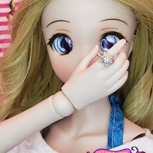 May include: A doll with blonde hair and blue eyes is wearing a white shirt and a blue and white patterned scarf. The doll is wearing a silver ring with a turtle design.