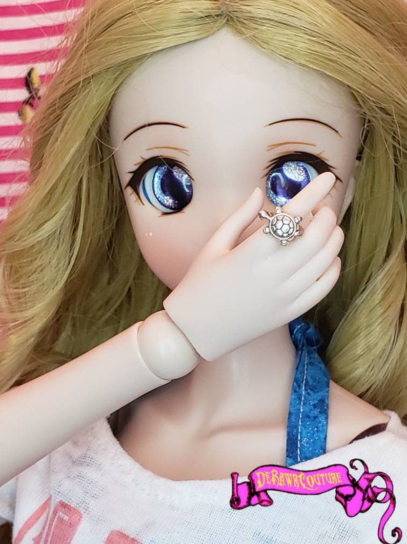 May include: A doll with blonde hair and blue eyes is wearing a white shirt and a blue and white patterned scarf. The doll is wearing a silver ring with a turtle design.