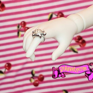 May include: A silver turtle ring with a detailed shell design. The ring is worn on a finger of a doll's hand.