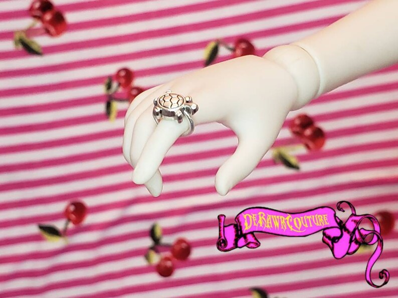 May include: A silver turtle ring with a detailed shell design. The ring is worn on a finger of a doll's hand.