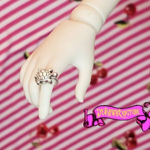 May include: A silver turtle ring with a textured shell design. The ring is worn on a hand against a pink and white striped background.
