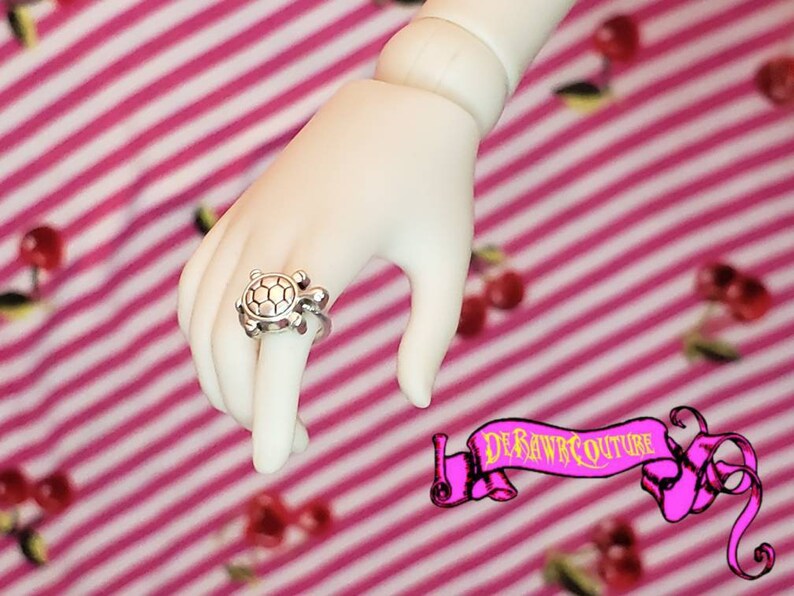 May include: A silver turtle ring with a textured shell design. The ring is worn on a hand against a pink and white striped background.