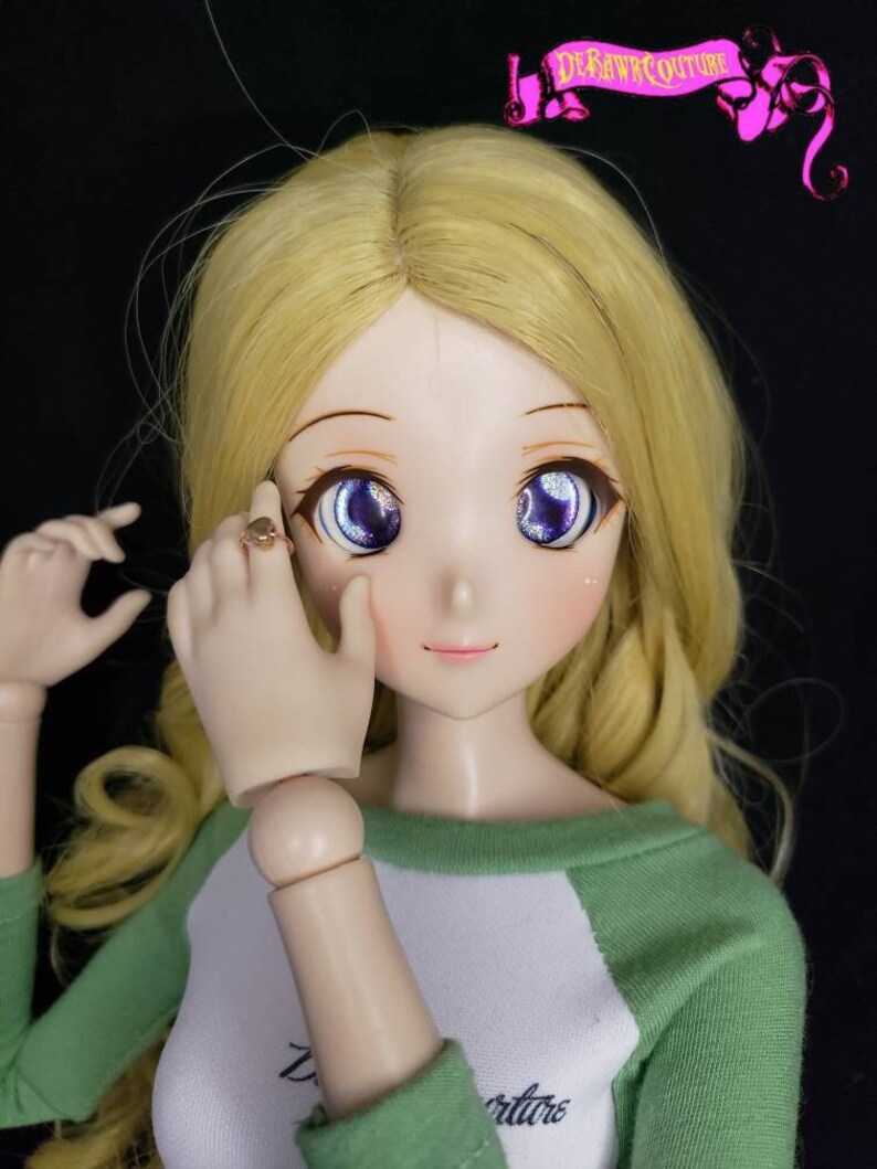 May include: A doll with long blonde hair wearing a green and white shirt. The doll has blue eyes with glitter and is wearing a gold ring on her finger.