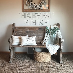 Puede incluir: Un banco de madera con una manta blanca y una almohada con la palabra "retreat" en ella. El banco est&aacute; frente a una pared con las palabras "HARVEST FINISH" escritas en ella.