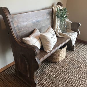 Puede incluir: Un banco de madera r&uacute;stico con respaldo alto y detalles tallados. El banco est&aacute; decorado con dos almohadas, una manta y un jarr&oacute;n con vegetaci&oacute;n. El banco est&aacute; sobre una alfombra tejida.