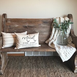 Puede incluir: Un banco de madera r&uacute;stico con una manta blanca y un jarr&oacute;n con follaje verde. Dos almohadas est&aacute;n en el banco, una con un patr&oacute;n de rayas y la otra con la palabra "retreat" y su definici&oacute;n.