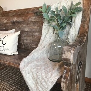 Puede incluir: Un banco de madera r&uacute;stico con un acabado blanqueado, que presenta una almohada blanca con la palabra "retreat" y su definici&oacute;n. Una manta de punto blanca y un jarr&oacute;n de vidrio con follaje verde se colocan en el banco.