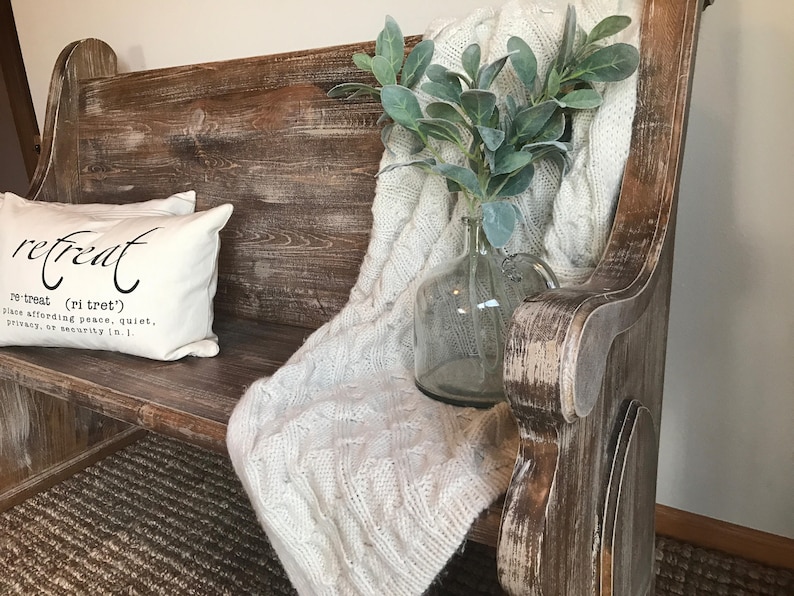 Puede incluir: Un banco de madera r&uacute;stico con un acabado blanqueado, que presenta una almohada blanca con la palabra "retreat" y su definici&oacute;n. Una manta de punto blanca y un jarr&oacute;n de vidrio con follaje verde se colocan en el banco.