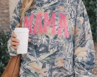 May include: A camouflage-patterned sweatshirt with the word "MAMA" in bright pink block letters across the front. The sweatshirt has a crew neck and long sleeves. The person is wearing blue jeans and carrying a white coffee cup.