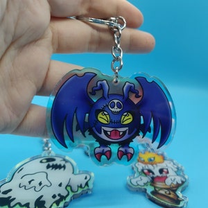 Può includere: Un portachiavi in acrilico iridescente blu e viola con un mostro dei cartoni animati con ali, un teschio e una bocca cucita. Il portachiavi è attaccato a una catena di metallo argentata.