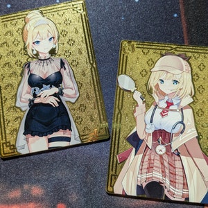 May include: Two gold-colored cards with illustrations of anime characters. The card on the left features a woman with blonde hair wearing a black dress and a white lace overlay. The card on the right features a woman with blonde hair wearing a white shirt, a brown coat, and a red and white plaid skirt. She is holding a magnifying glass in her right hand.