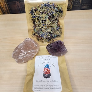 May include: A clear plastic bag filled with dried herbs and flowers, next to a brown paper bag with the text "Witches Brew Tea." Two gemstones, one pink and one purple, are also present. The tea is labeled as "Color Changing herbal tea."