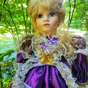 May include: A porcelain doll with blonde hair and blue eyes, wearing a purple dress with gold and white lace trim. The doll has a gold flower accent on the bodice and is set against a backdrop of green foliage.