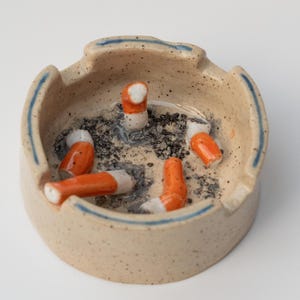 以下が含まれることがあります： ベージュ色の陶器の灰皿。まだら模様の仕上がりで、青い縁取りがある。オレンジと白のフィルターが付いたタバコの吸い殻がいくつかあり、灰に囲まれている。