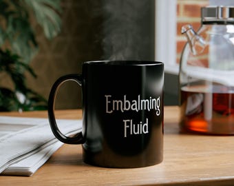 May include: A black ceramic mug with the text "Embalming Fluid" in white lettering. The mug is filled with a steaming beverage.
