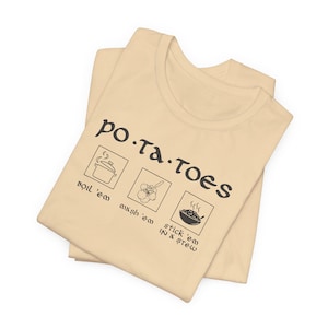 May include: A beige t-shirt with the text "po.ta.toes" in black lettering. The shirt has three black and white illustrations of different ways to prepare potatoes: boil 'em, mash 'em, and stick 'em in a stew.