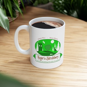 May include: White ceramic mug filled with dark coffee, featuring a green and white graphic of a castle, trees, and a rabbit. The mug has the text "Roger's Shrubbery" and "Always Nice - Never Showy".