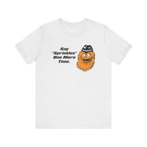 Könnte beinhalten: Ein hellgraues T-Shirt mit einem orangefarbenen Monster-Cartoon, das einen schwarzen Hut trägt. Das Monster hat einen überraschten Ausdruck und der Text "Say "Sprinkles" One More Time." ist unter dem Monster gedruckt.