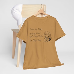 Op de afbeelding: Een mosterdgeel t-shirt met een zwarte afbeelding van een cartoonfiguur en de tekst "This is Sam Nothing Ever Get's Sam Down Be Like Sam".