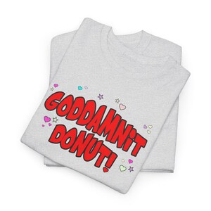 May include: Two light gray t-shirts are stacked. The top shirt has the words "GODDAMNIT DONUT!" in large, red, bubble letters. Small hearts and stars in various colors surround the text.