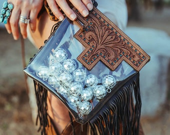 May include: A clear plastic purse with a brown leather flap and fringe detail. The flap has a tooled design and is decorated with turquoise studs. The purse is filled with silver disco balls.