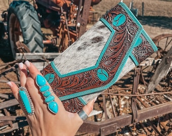 May include: A white and brown leather wallet with turquoise accents and floral tooling. The wallet is held in a hand with turquoise rings and bracelets.