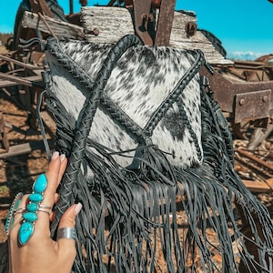 Puede incluir: Un bolso bandolera de piel de vaca blanco y negro con flecos negros y una correa de cuero trenzada. El bolso está colgado de una estructura metálica oxidada.