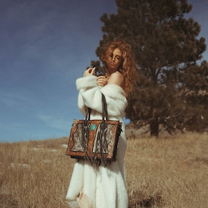 May include: A woman wearing a white fur coat carries a brown leather tote bag with turquoise accents and fringe. The bag features a distinctive design with intricate details. The woman is standing in a field of long grass with a large tree behind her.