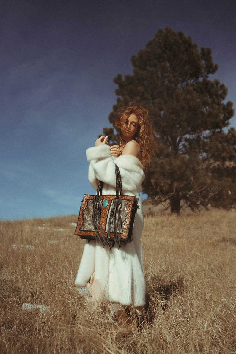 May include: A woman wearing a white fur coat carries a brown leather tote bag with turquoise accents and fringe. The bag features a distinctive design with intricate details. The woman is standing in a field of long grass with a large tree behind her.