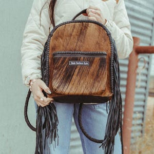 May include: A brown and black cowhide backpack featuring a front pocket with the text Haute Southern Hyde, a braided leather trim along the edges, and long black leather fringe tassels hanging from the sides and the base of the bag.