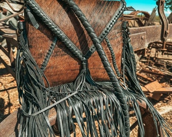 Puede incluir: Un bolso de mano de piel de vaca marrón y negro con una larga correa trenzada negra y flecos negros. El bolso está colgado de una estructura metálica oxidada.