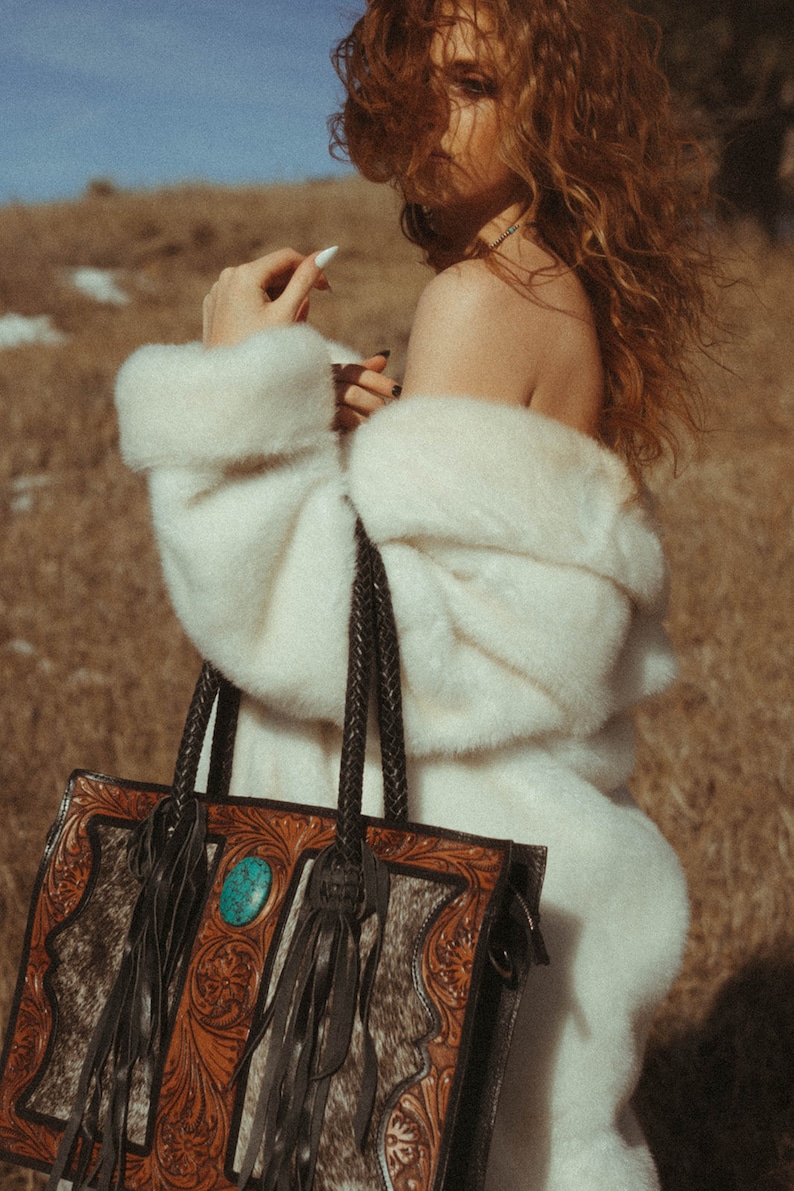 May include: A brown leather tote bag with a turquoise stone and fringe detail. The bag has a braided strap and is decorated with a floral pattern.