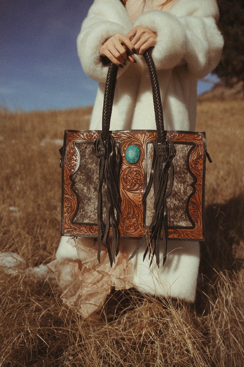 May include: A brown leather tote bag with intricate floral tooling and a turquoise stone accent. The bag has black fringe and a cowhide panel.